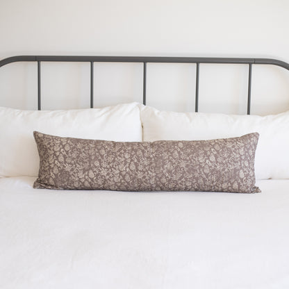 Taupe lumbar pillow with cream floral damask pattern displayed on white bedding against metal headboard.