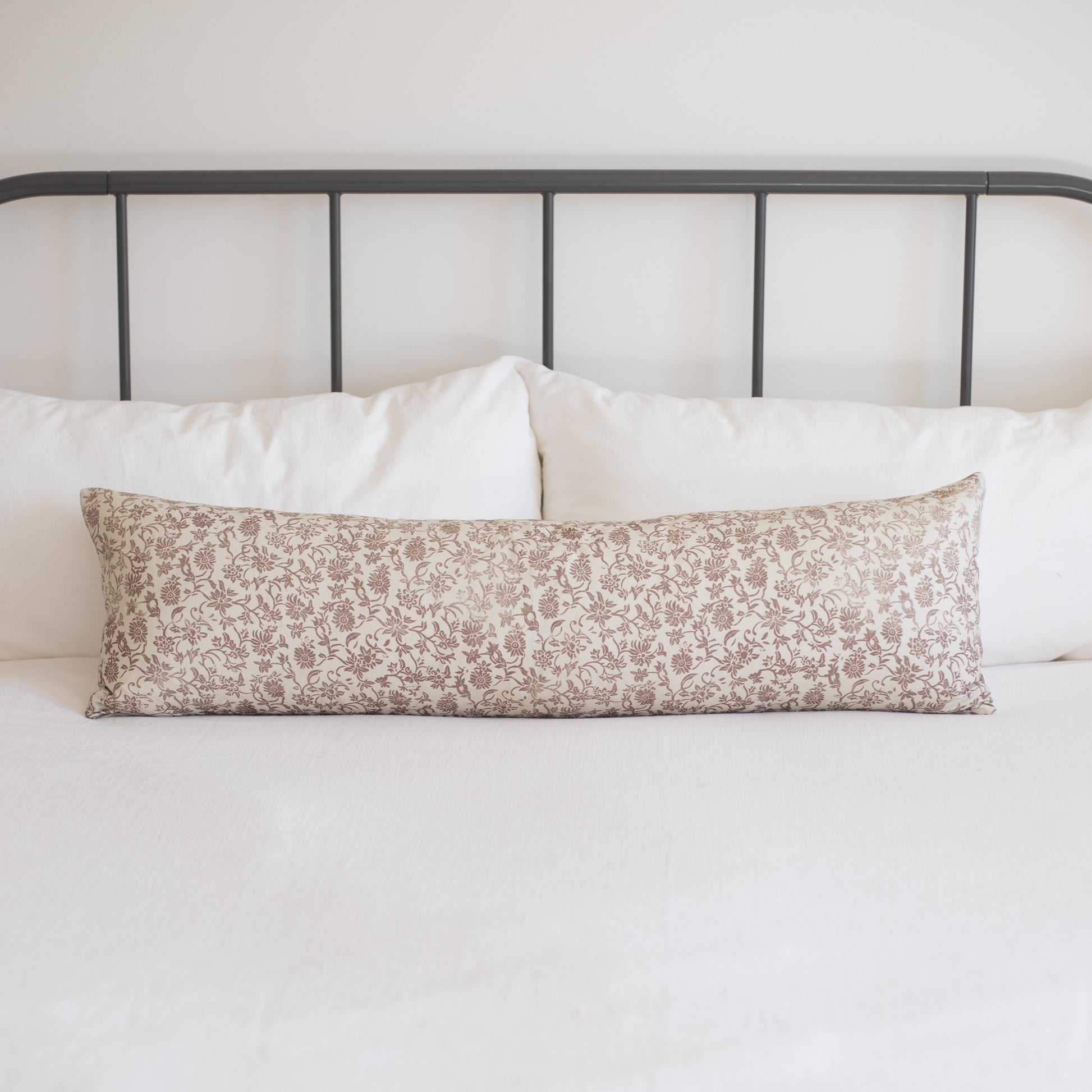 Rectangular lumbar pillow with beige floral pattern on white bedding against metal headboard.