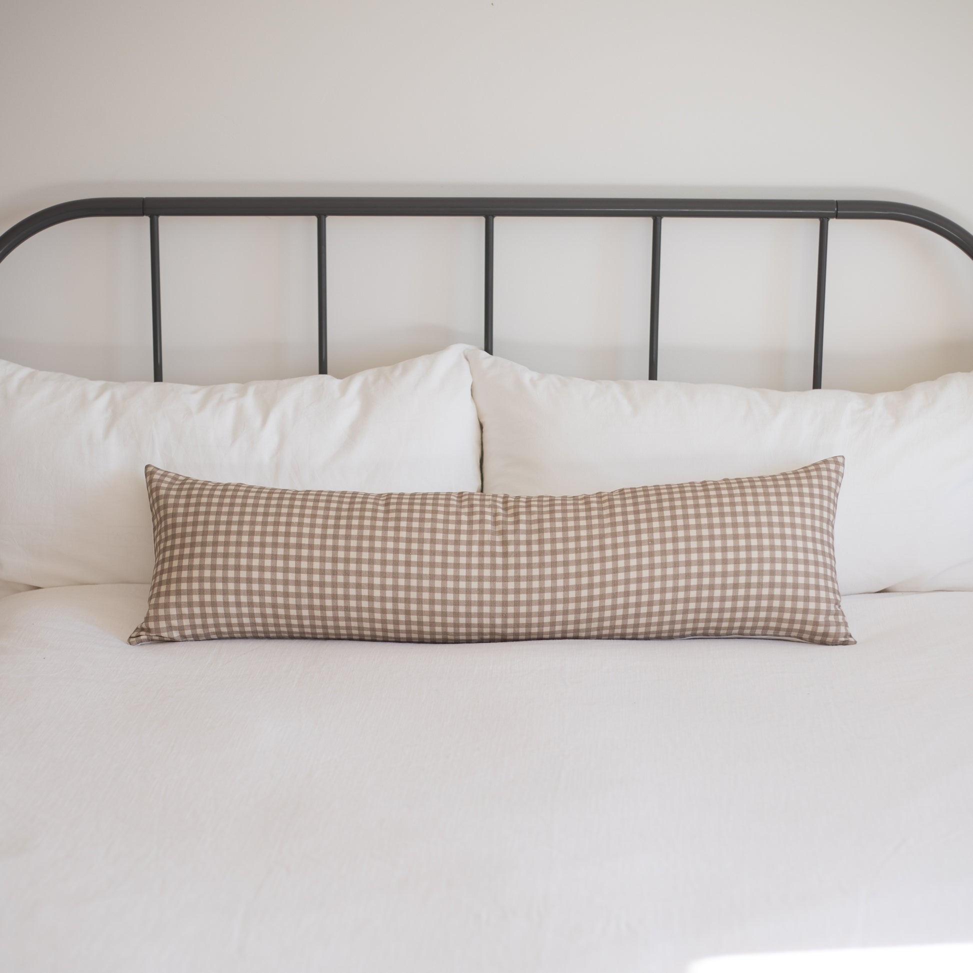 Tan and white gingham lumbar pillow displayed on a bed with white pillows and black metal headboard.