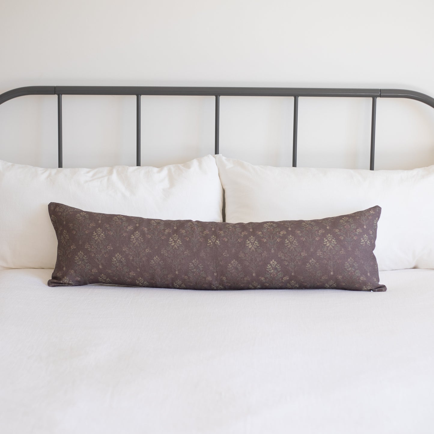 Burgundy linen lumbar pillow with small floral pattern, displayed on bed with white pillows and metal headboard.