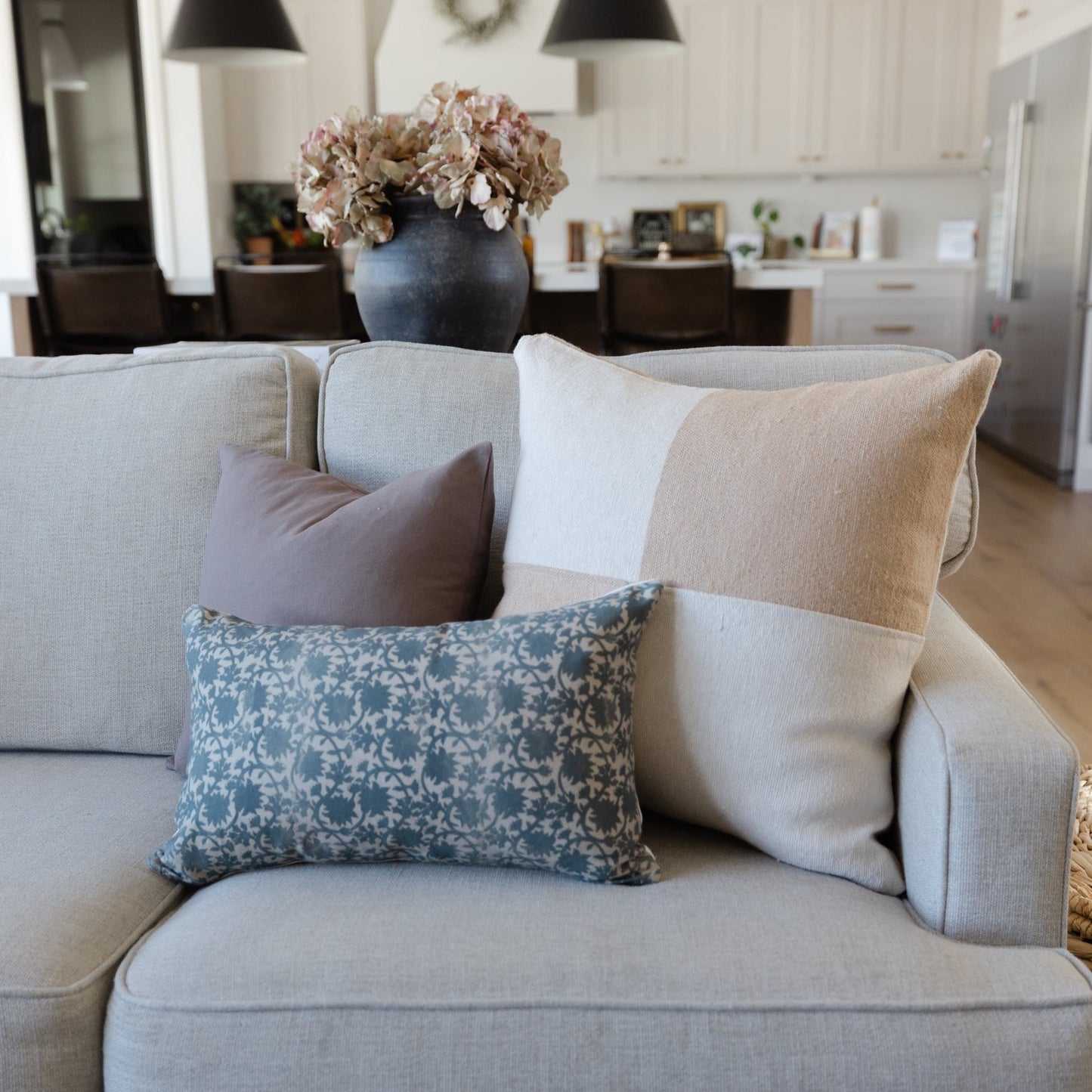 Tan and cream textured throw pillows arranged on a light gray upholstered sofa with blue floral pattern accent pillow.