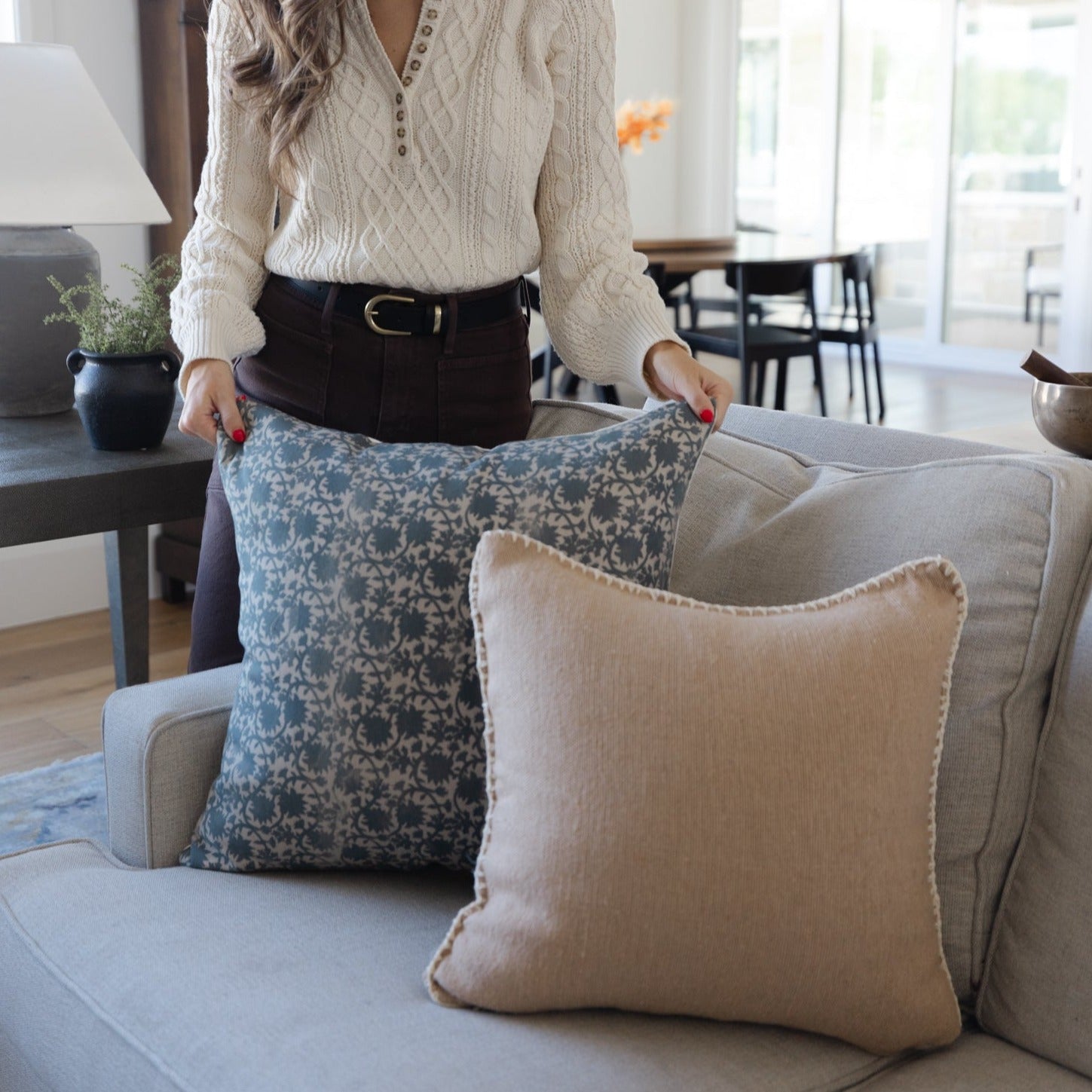 Woman holding blue damask patterned pillow and cream textured pillow on beige sofa.