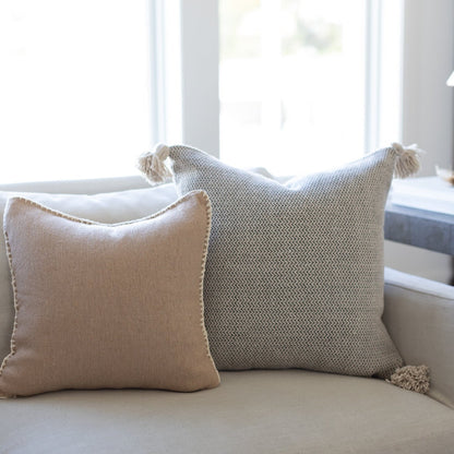 Cream tweed throw pillow with textured weave pattern and tassel corners displayed on a light sofa near a window.