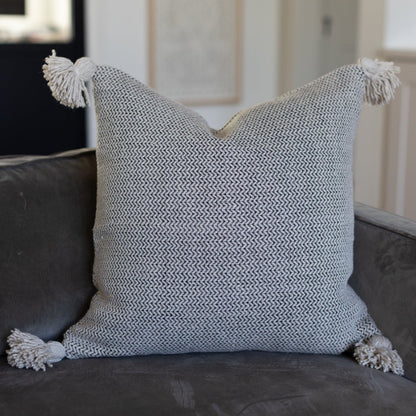Cream and gray tweed pillow with corner tassels displayed on a dark sofa.