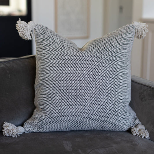 Cream and gray tweed pillow with corner tassels displayed on a dark sofa.