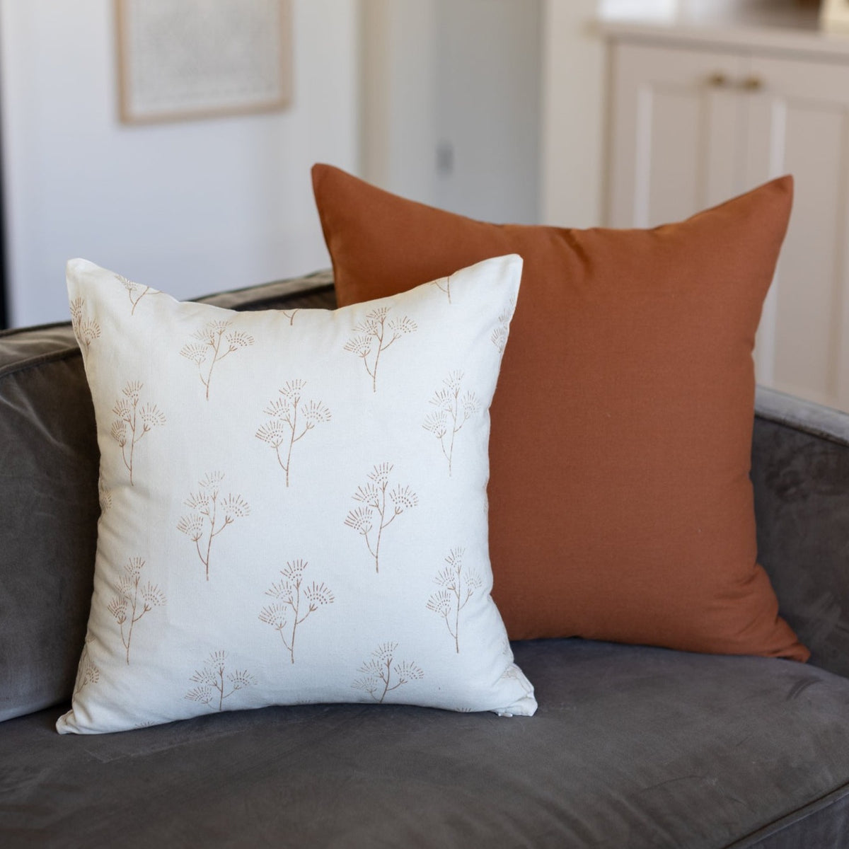 Cream pillow with tan thistle print and rust solid pillow on gray sofa.