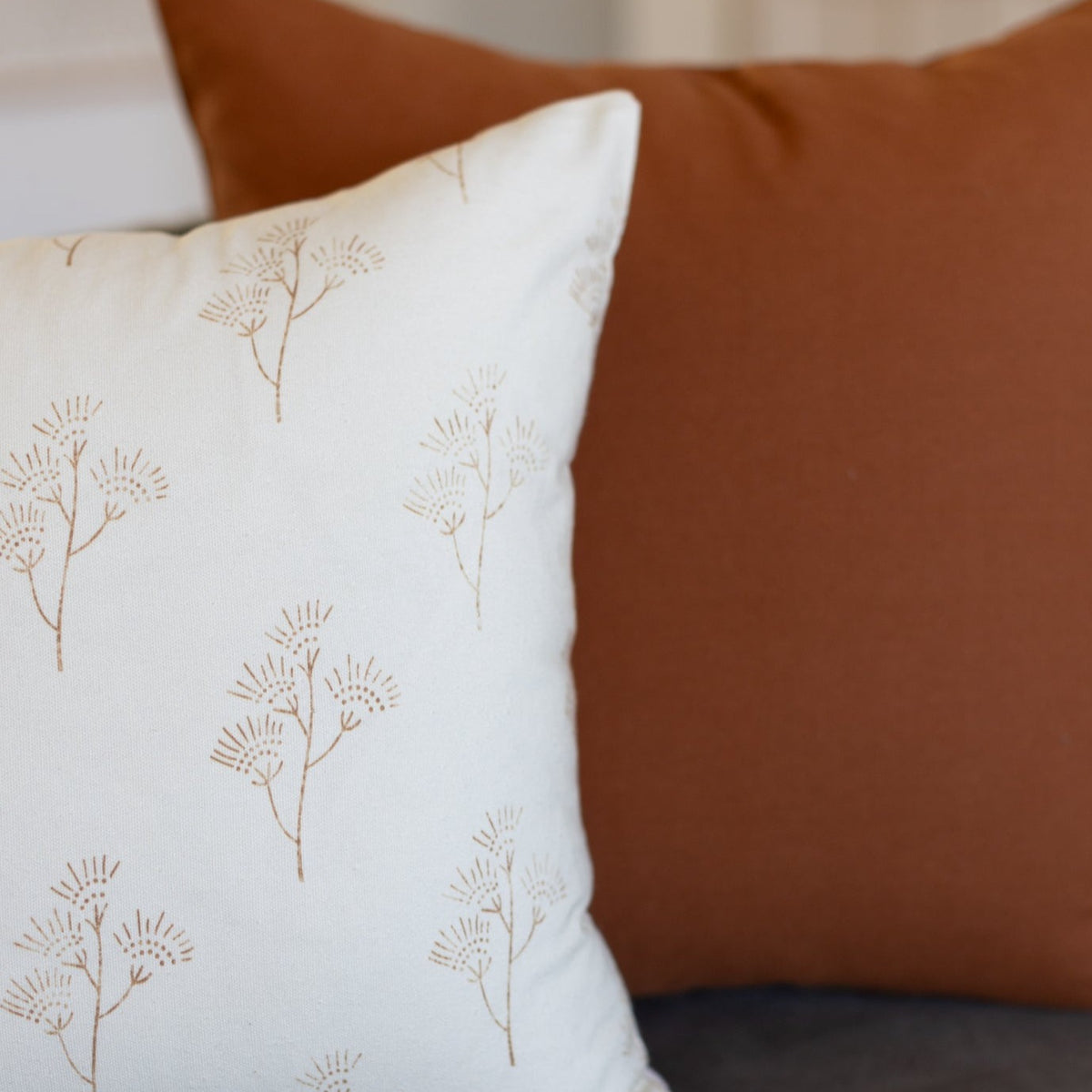 White pillow with tan thistle plant print, displayed next to solid brown pillow.