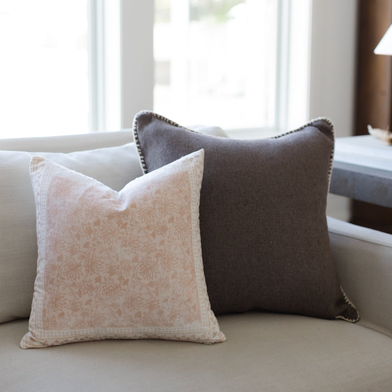 Warm charcoal throw pillow with nailhead trim displayed on a cream sofa next to a patterned cushion.