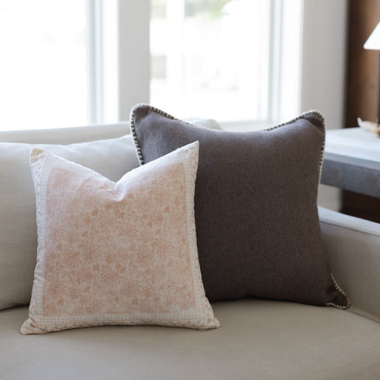 Two decorative pillows on a light sofa: one beige with damask pattern, one charcoal gray with nailhead trim.