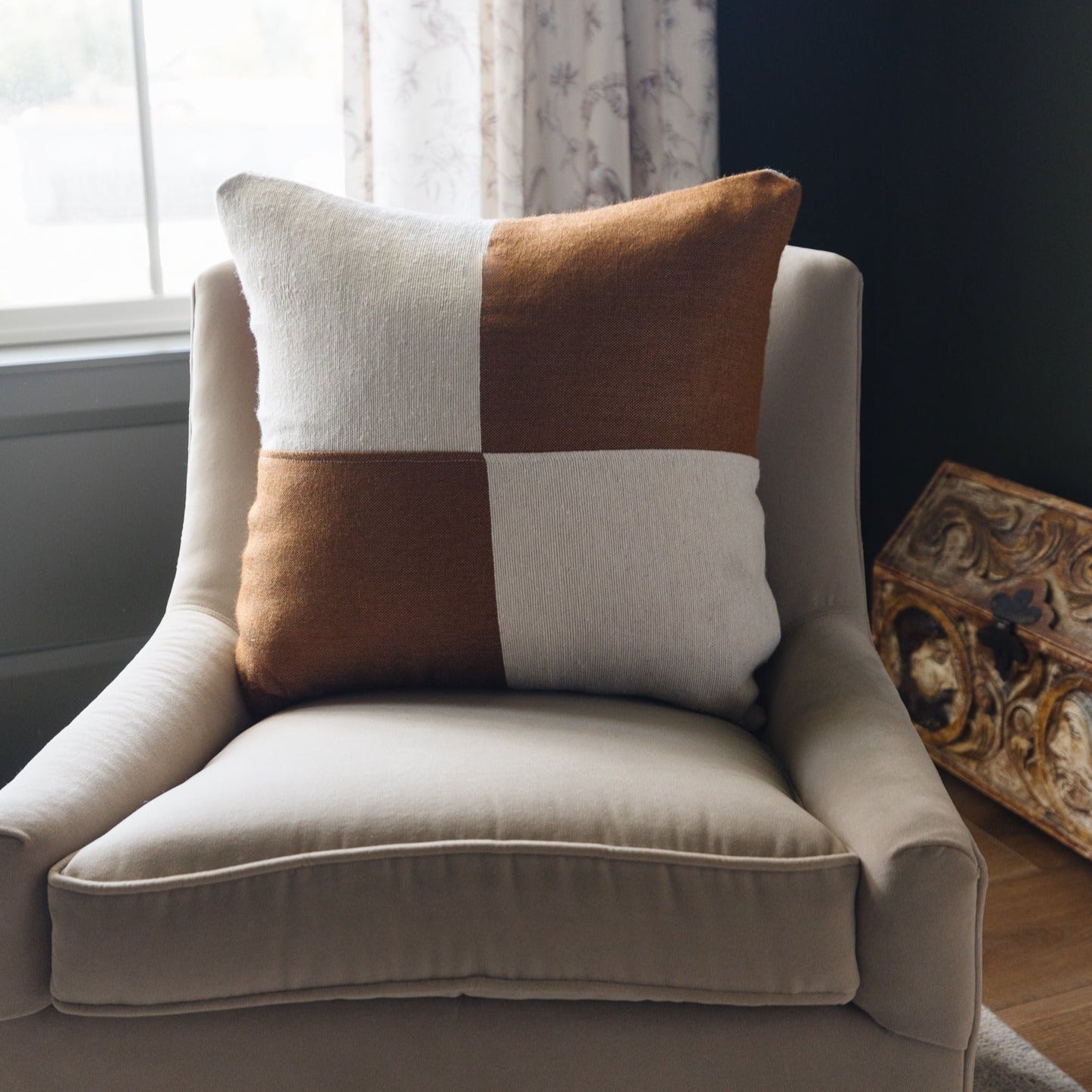 Square pillow with brown and cream checkered pattern on beige upholstered chair by window.