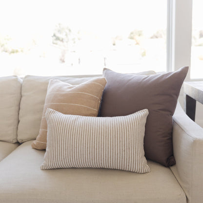 Three decorative pillows in mocha, tan, and cream tones arranged on a beige sofa by a window.