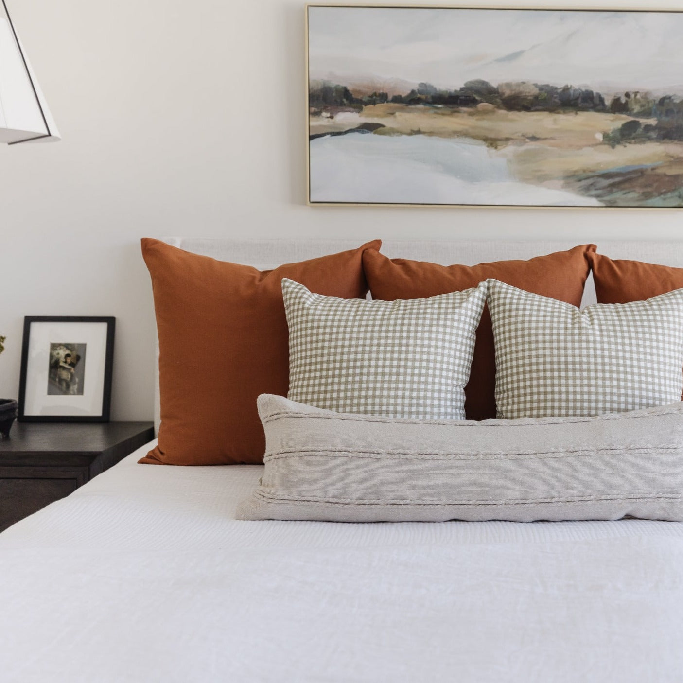 Two tan and white gingham patterned pillows on a bed with rust and cream pillows and white bedding.