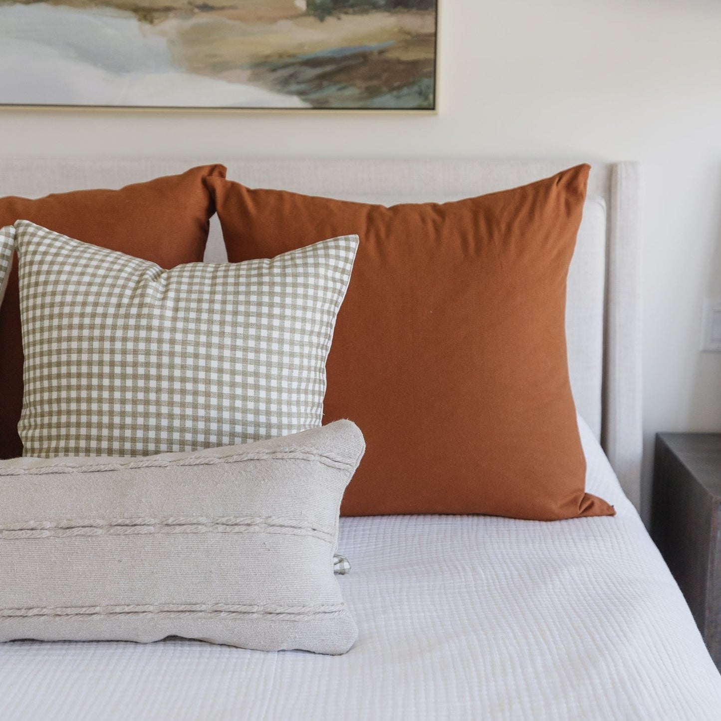 Tan and white gingham patterned throw pillow displayed on a bed with solid rust and beige pillows.