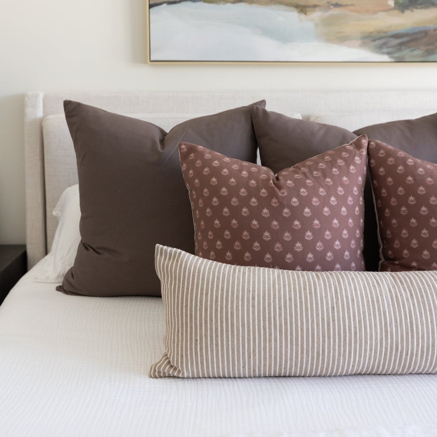 Mocha throw pillows in solid and patterned designs arranged on a bed with white bedding.