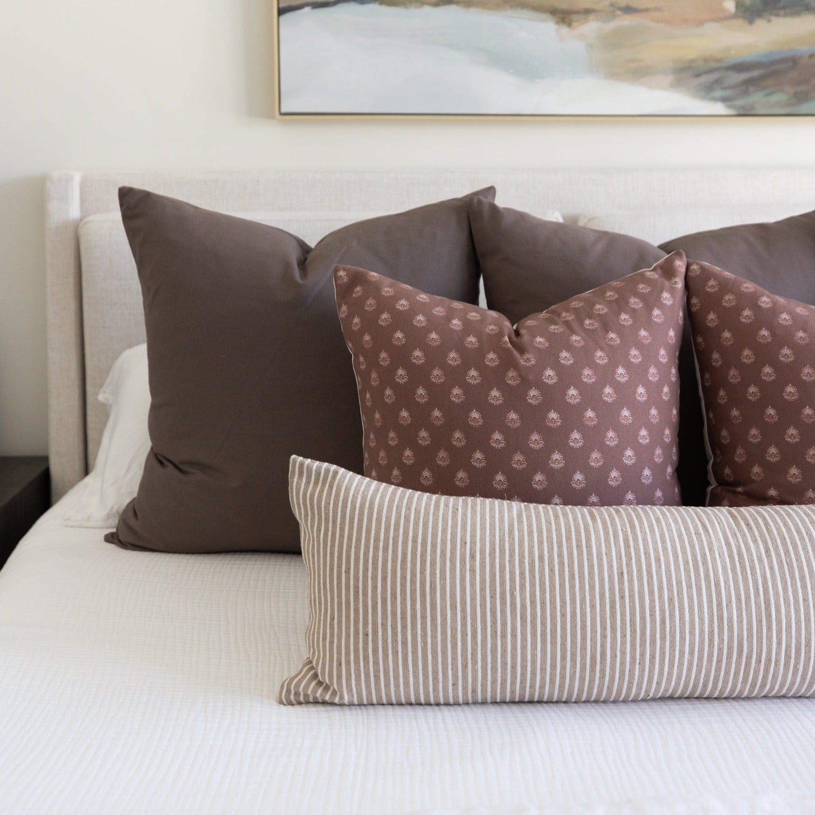 Mocha throw pillows in solid and patterned designs arranged on a bed with white bedding.