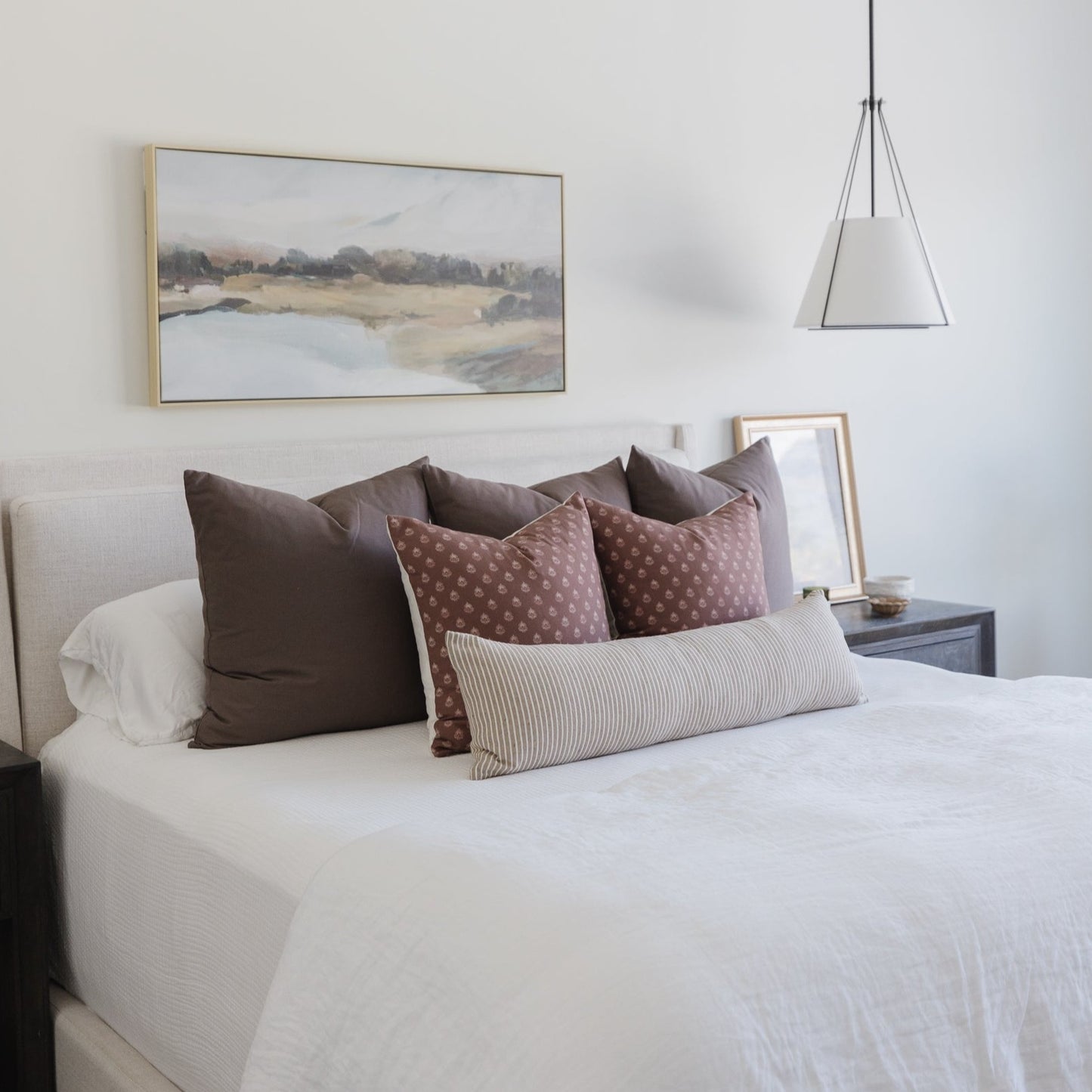Mocha-colored pillows arranged on a bed with white bedding, including solid and patterned cushions in various sizes.