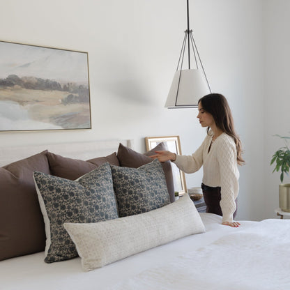 Rectangular lumbar pillow with cream textured fabric displayed on bed with patterned throw pillows.