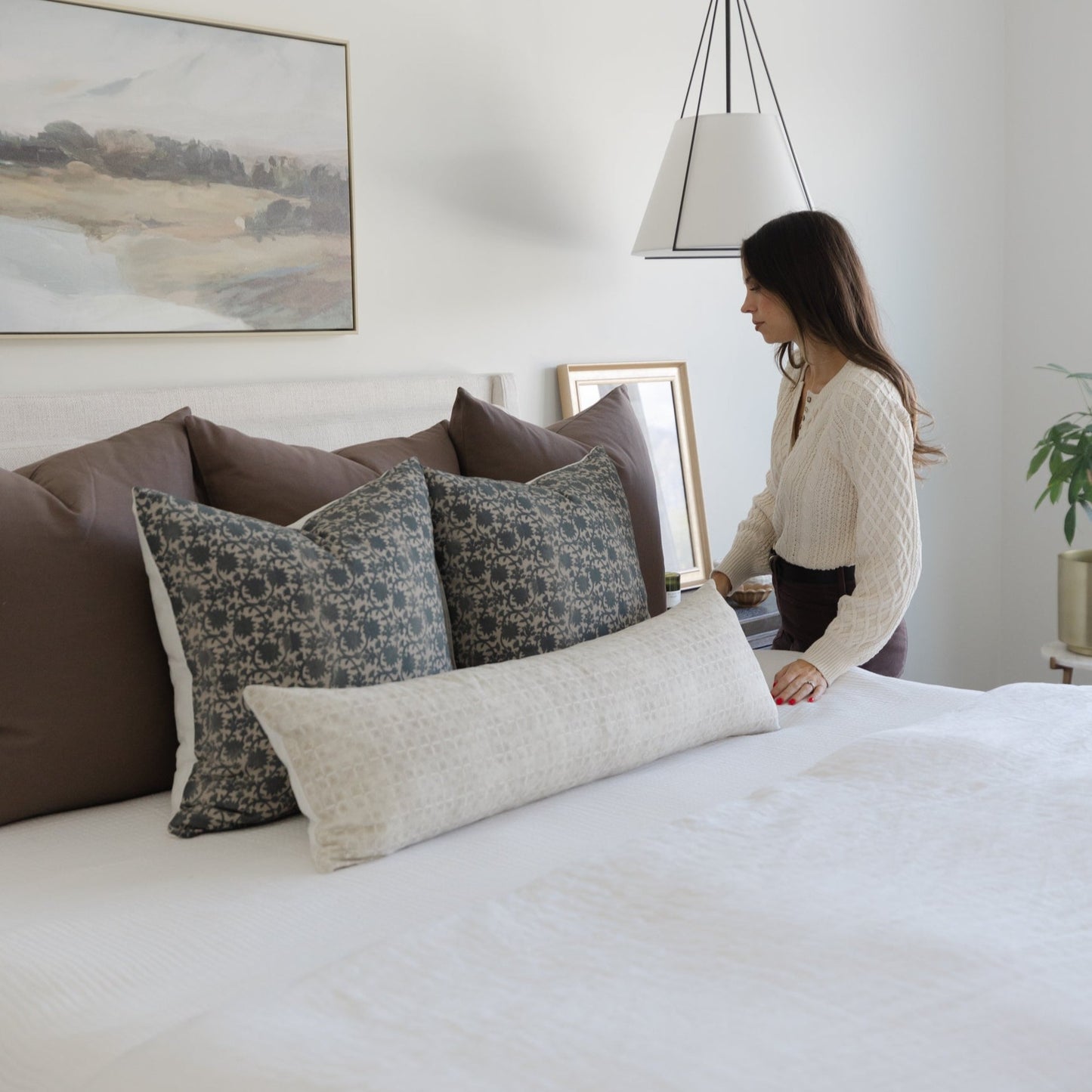 Mocha-colored and patterned throw pillows arranged on a bed with a woman standing beside them.