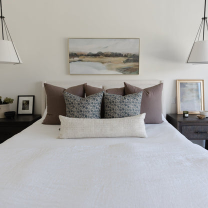Landscape artwork with muted tones showing a water and field scene, displayed above a bed with brown and patterned pillows.