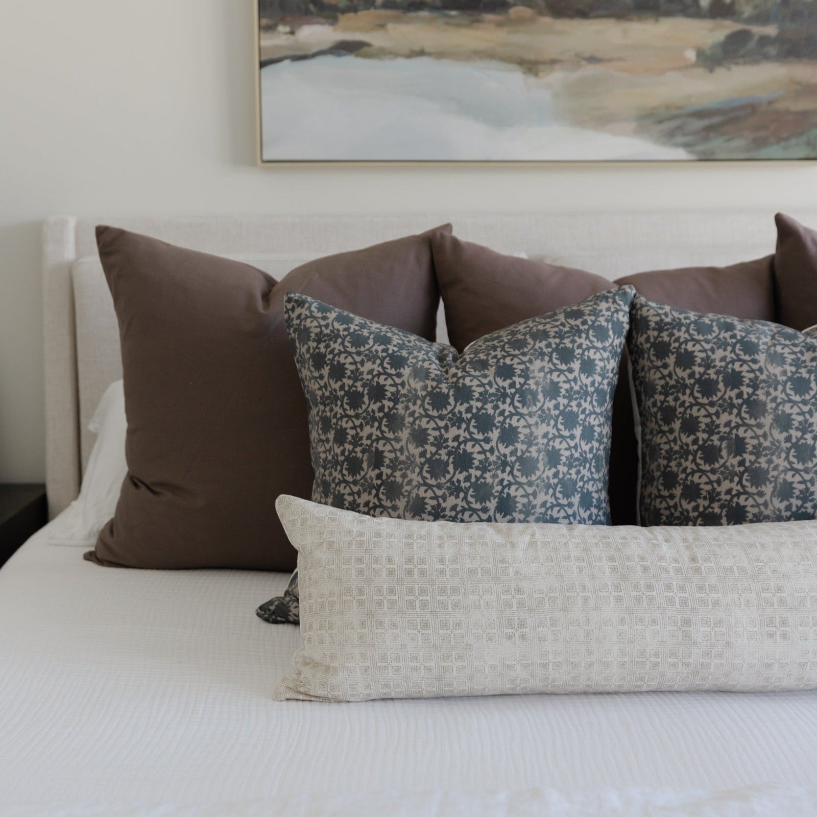 Mocha brown and patterned throw pillows arranged on a white bedding with a landscape artwork above.