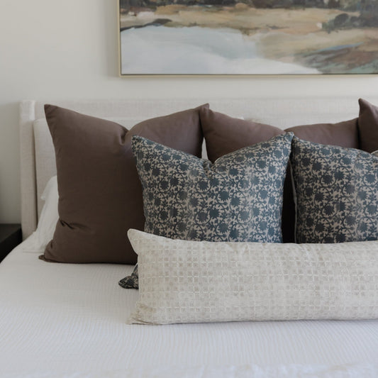 Mocha brown and patterned throw pillows arranged on a white bedding with a landscape artwork above.