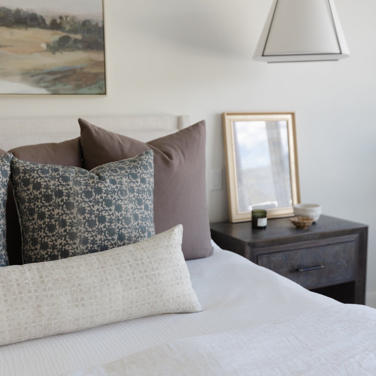 Bedroom styled with layered pillows in neutral tones and patterned fabrics on a white bedded bed.