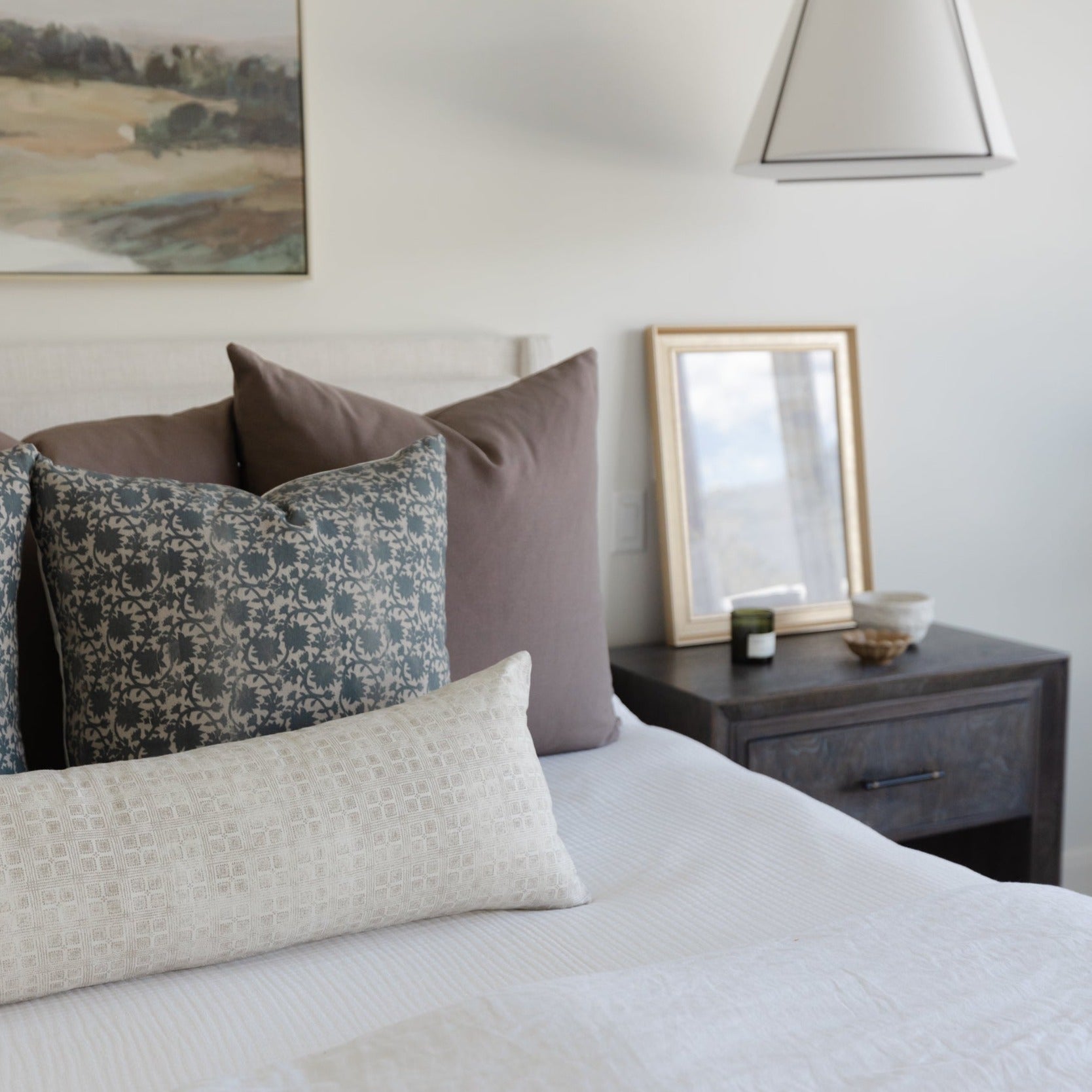 Bedroom styled with layered pillows in neutral tones and patterned fabrics on a white bedded bed.