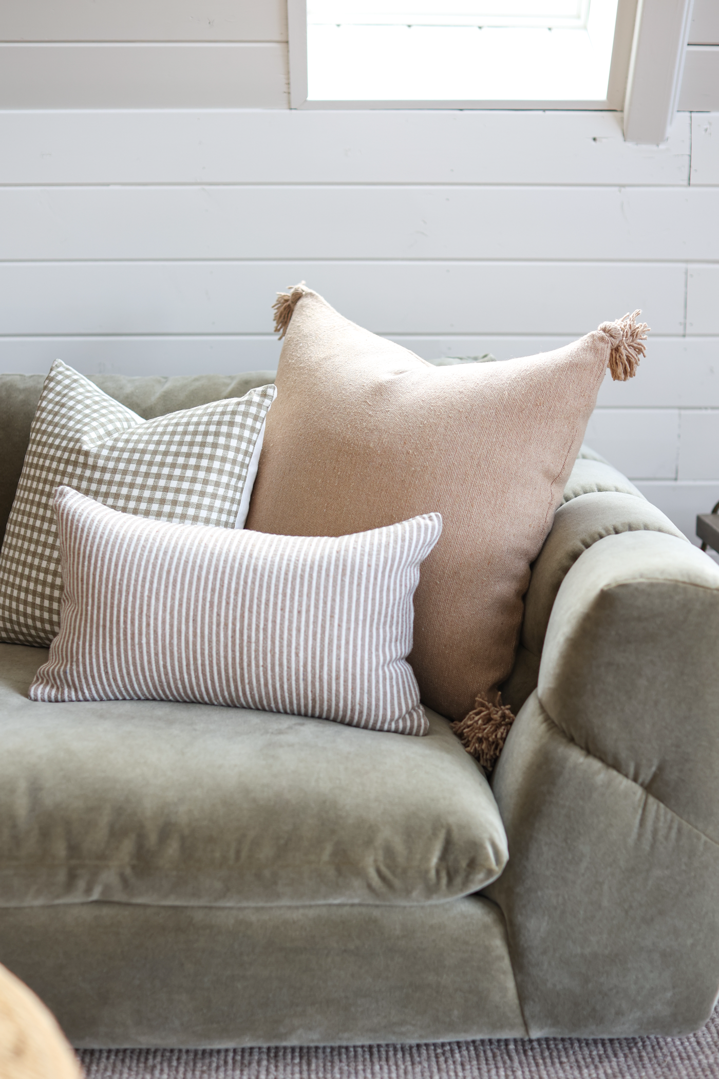 Tan lumbar pillow with tassel corners on gray upholstered chair with striped and checkered accent pillows.