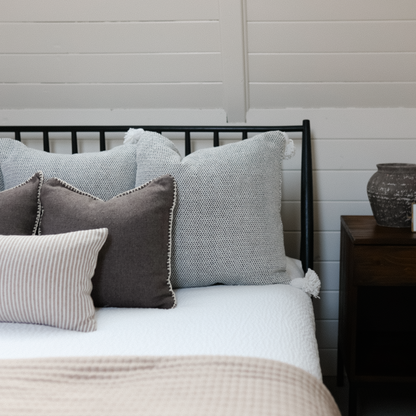 Warm charcoal throw pillow with nailhead trim displayed on bed with other decorative pillows.