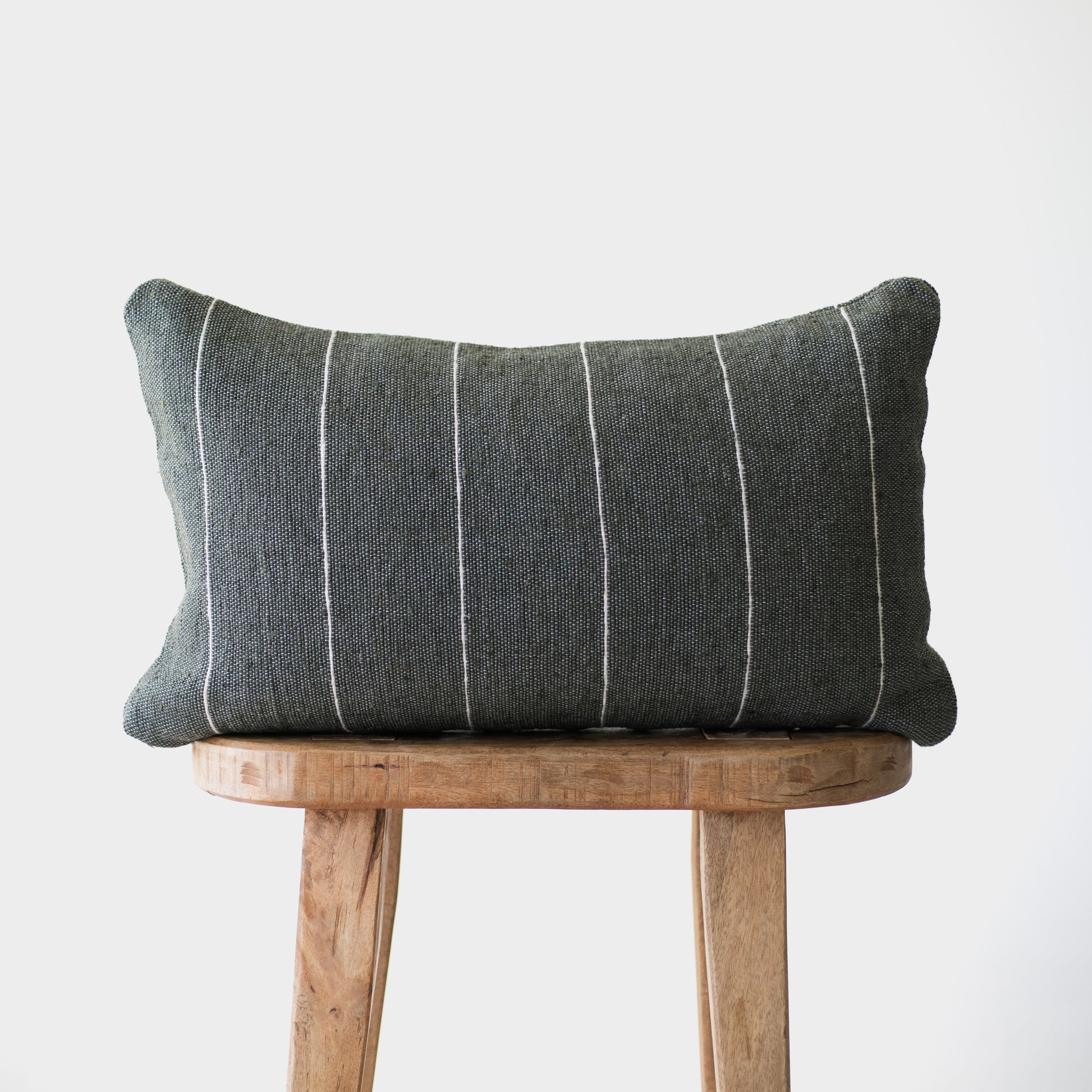 Moss-green lumbar pillow with vertical white stripes on wooden stool.
