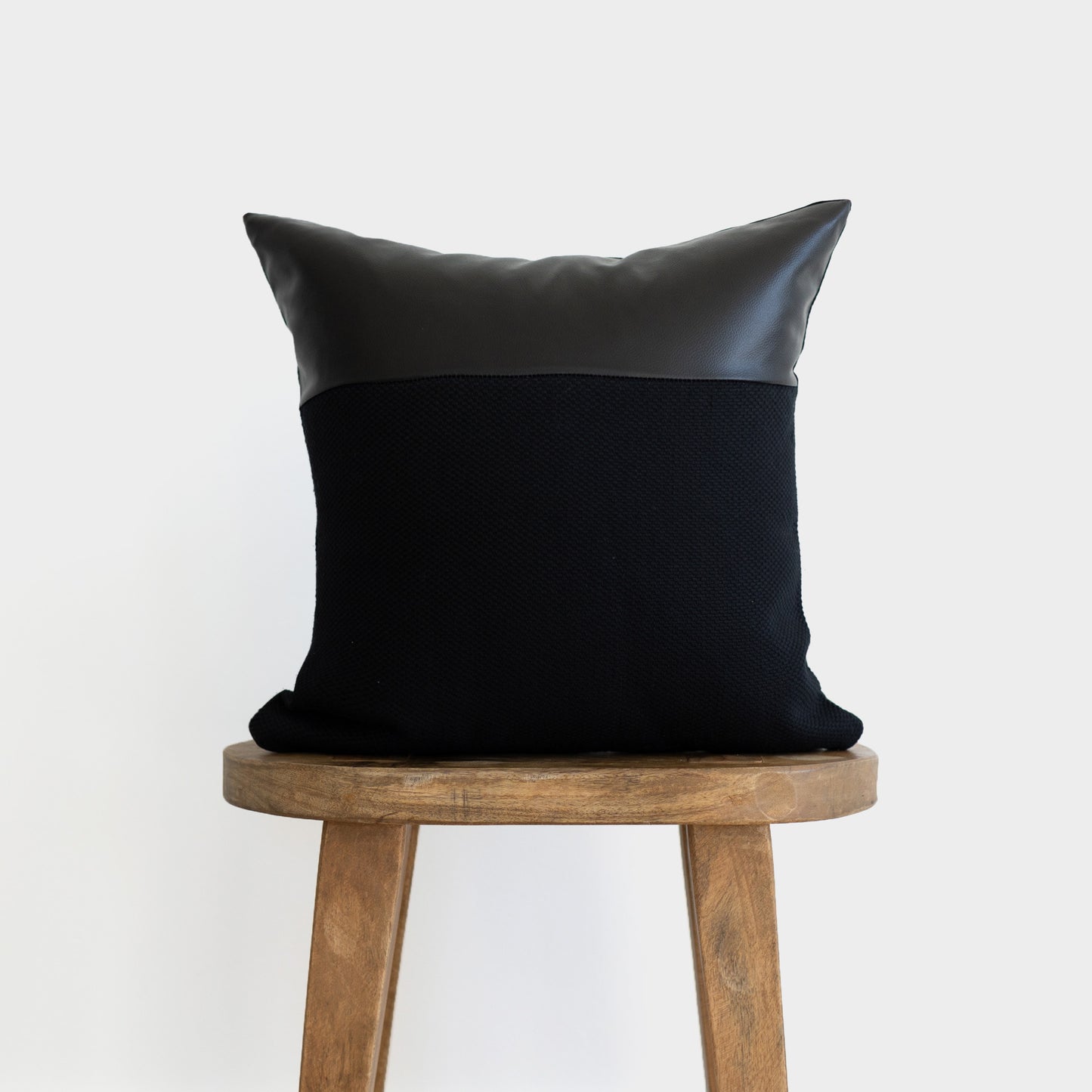 Black throw pillow with leather top and textured fabric body, displayed on wooden stool.