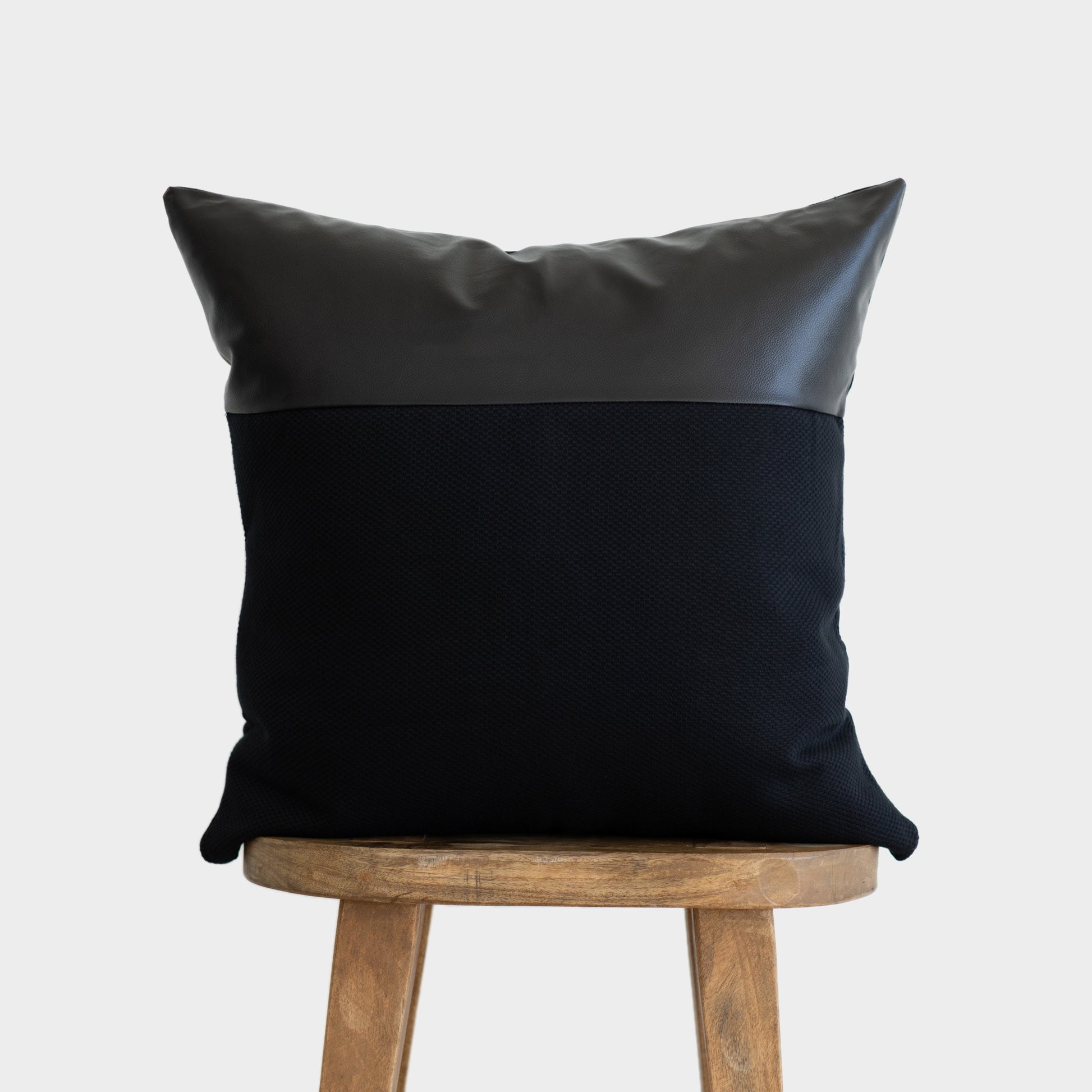 Black cushion with faux leather top panel and textured fabric body, displayed on wooden stool.