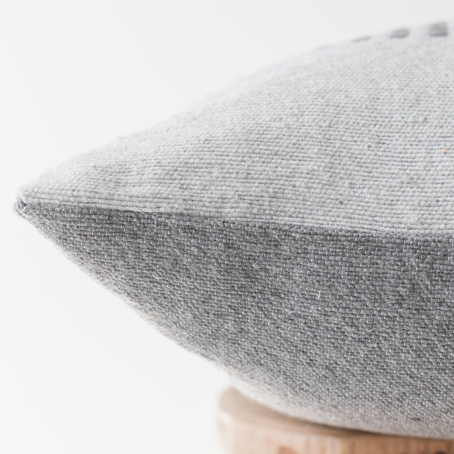 Gray woven pillow with piping detail on light wooden base, shown at angle.
