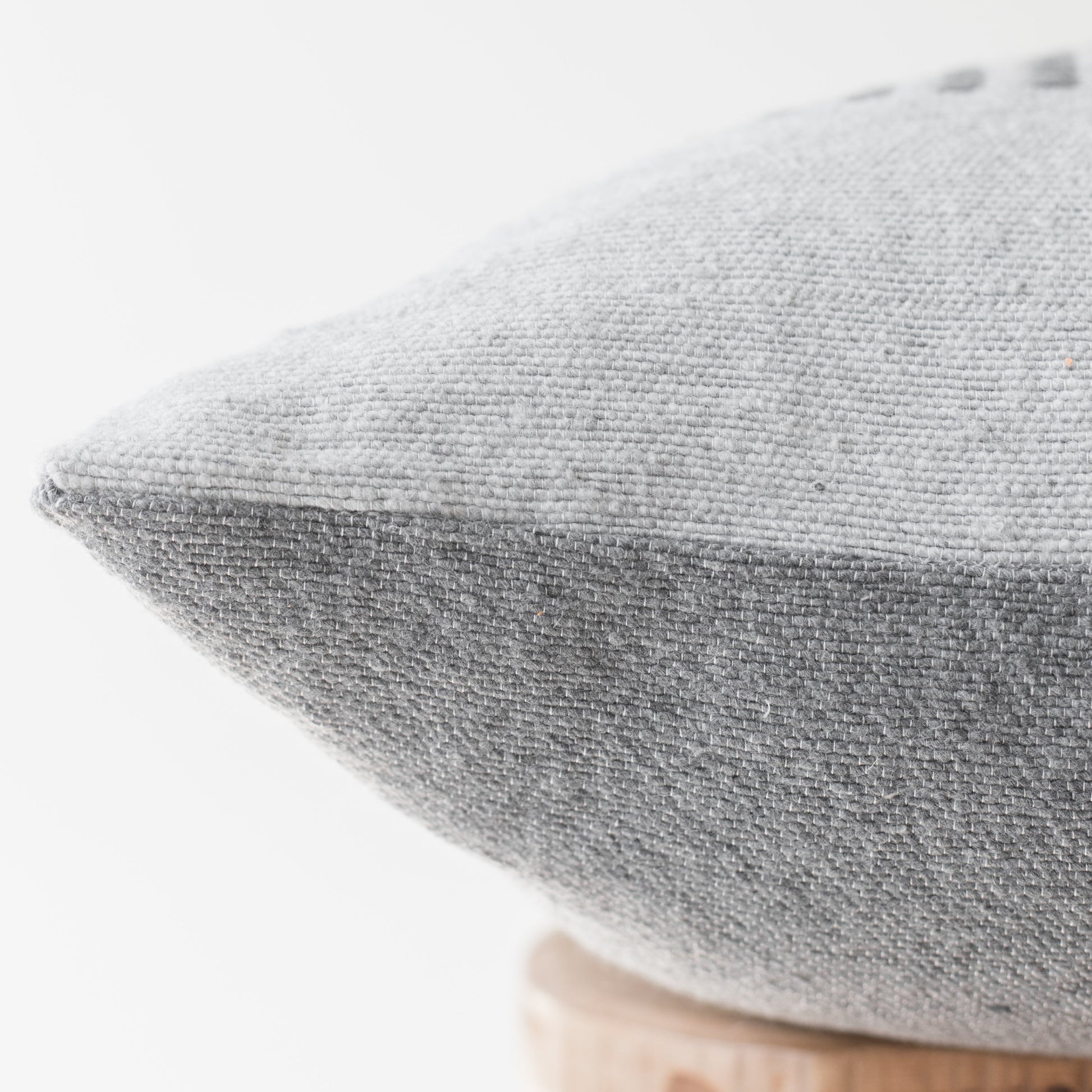 Gray woven pillow with piping detail on light wooden base, shown at angle.