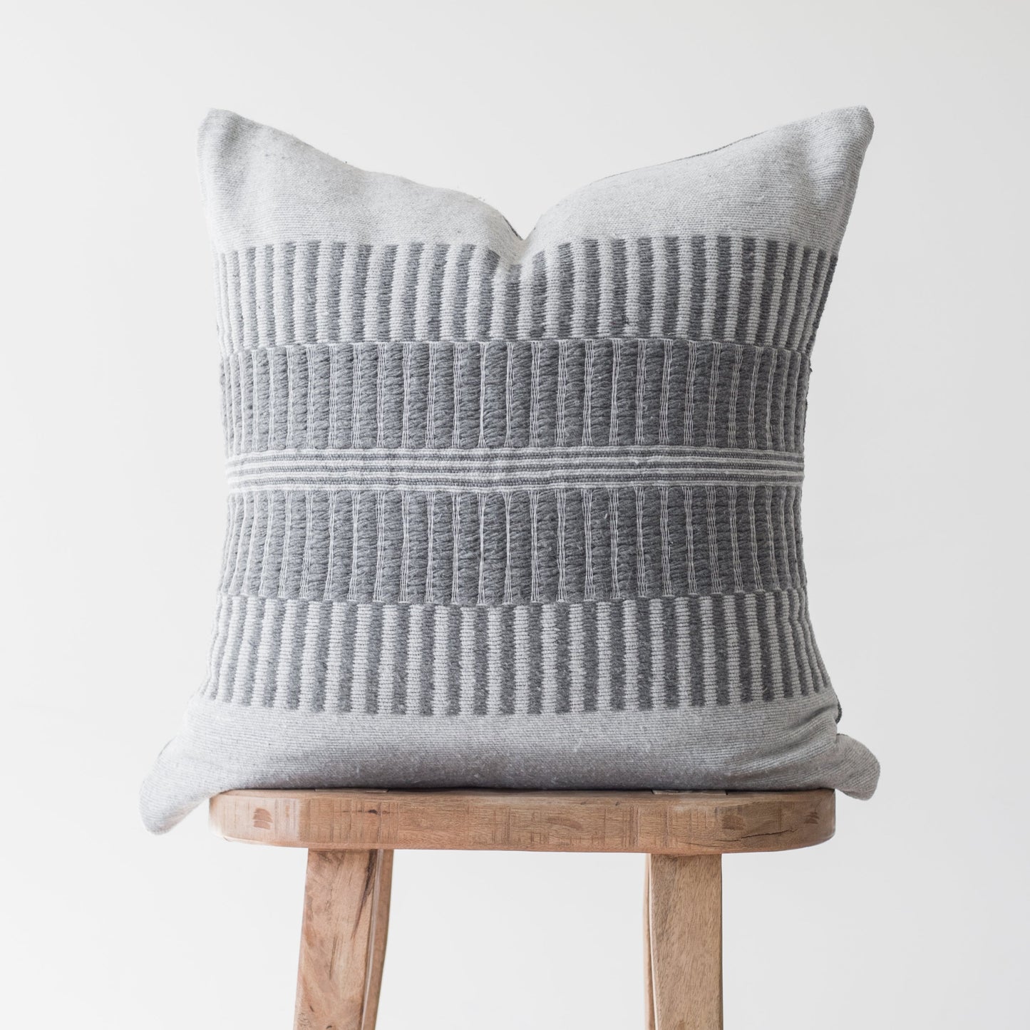 Light gray pillow with vertical ribbed texture and horizontal stripe detail, displayed on a wooden stool.