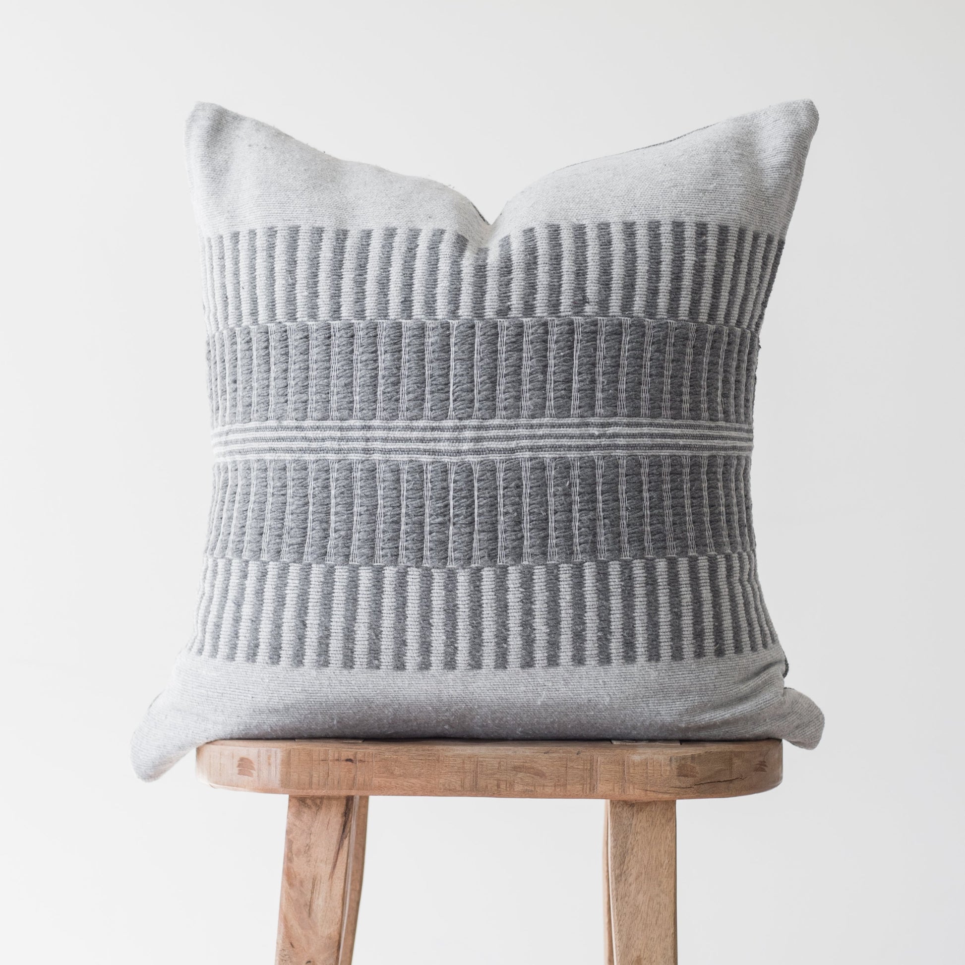 Light gray pillow with vertical ribbed texture and horizontal stripe detail, displayed on a wooden stool.