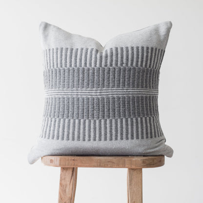Light gray pillow with vertical ribbed texture and horizontal stripe detail, displayed on a wooden stool.