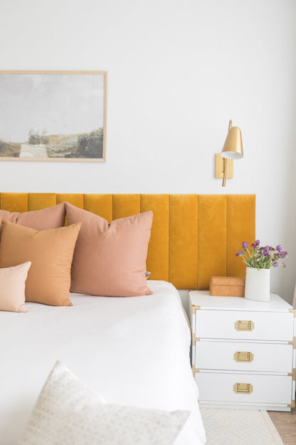 Dusty rose and tan square throw pillows arranged on a bed with yellow headboard and white bedding.