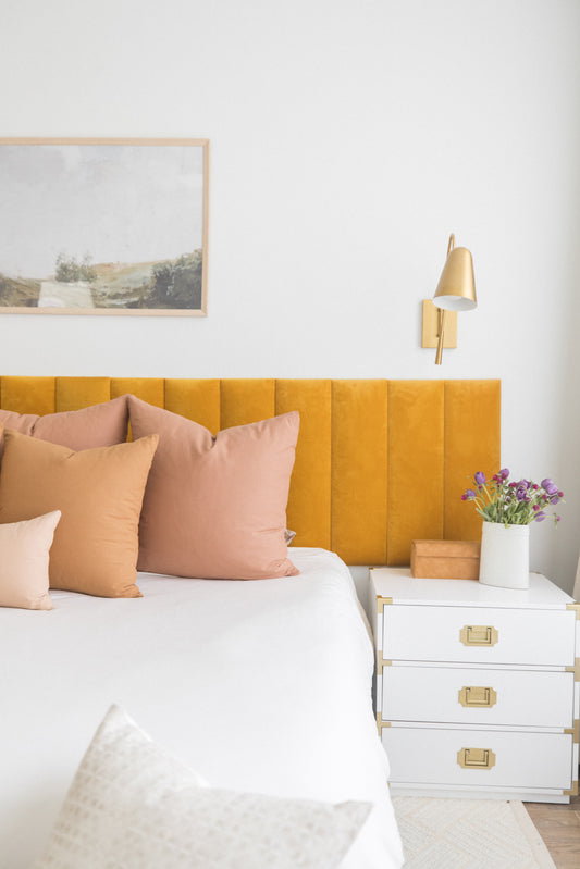 Dusty rose and tan square throw pillows arranged on a bed with yellow headboard and white bedding.