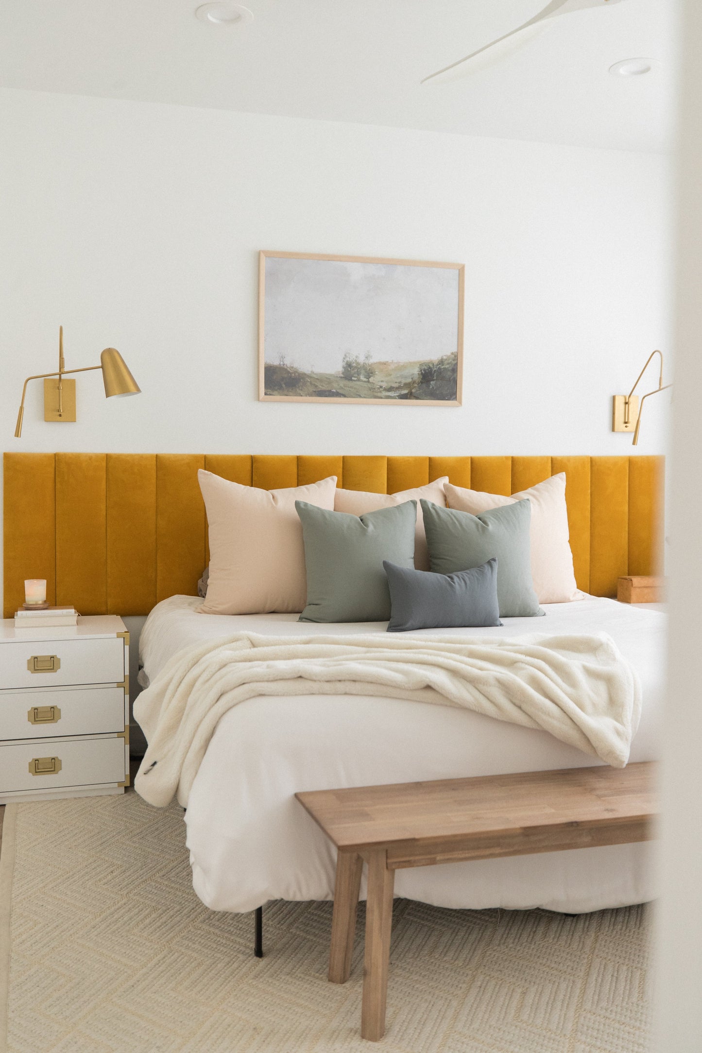 Bedroom with mustard yellow upholstered headboard, cream and gray pillows, white bedding, and decorative throw blanket.