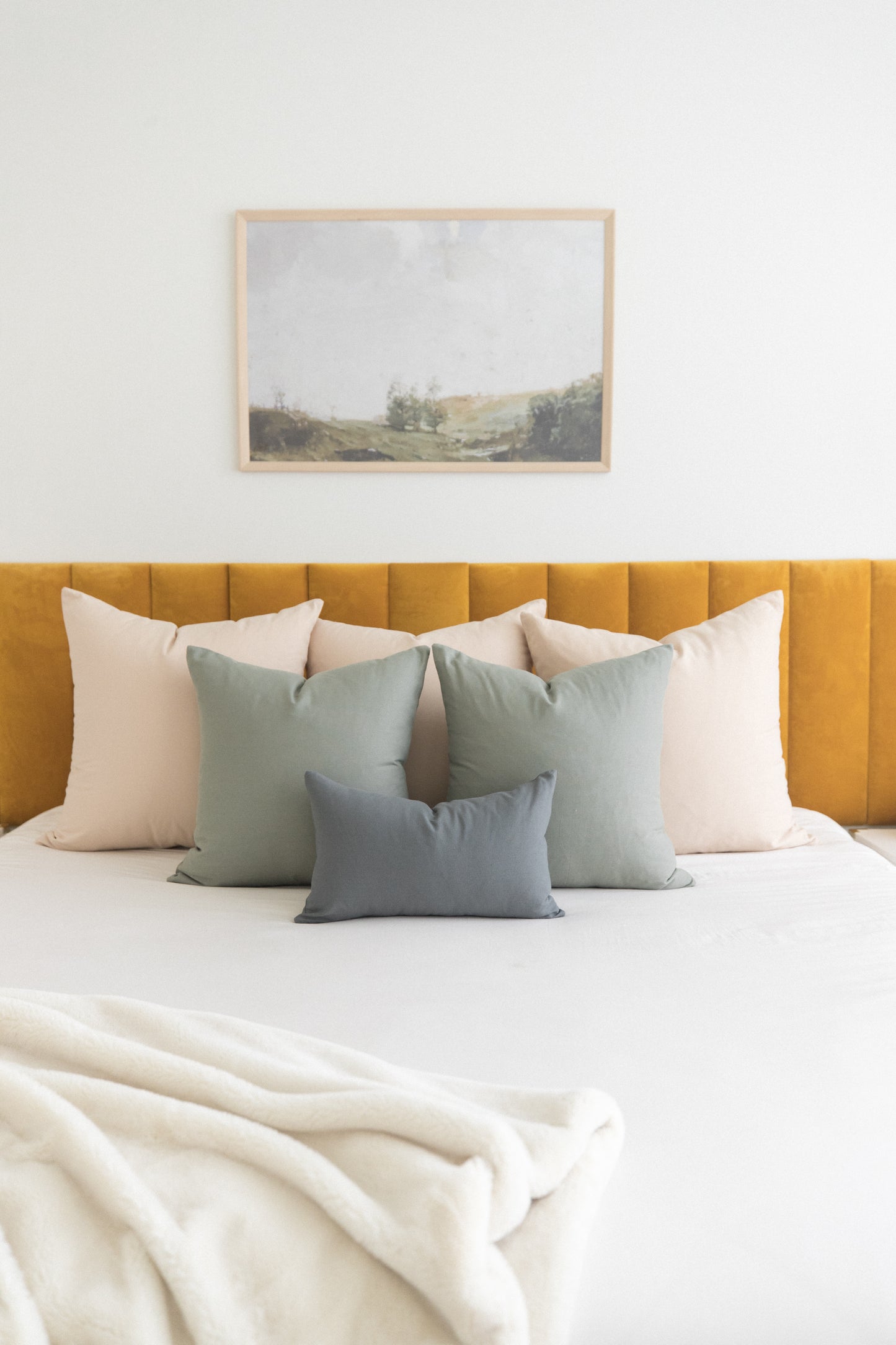 Beige and sage green throw pillows arranged on a bed with mustard yellow headboard and framed landscape artwork above.