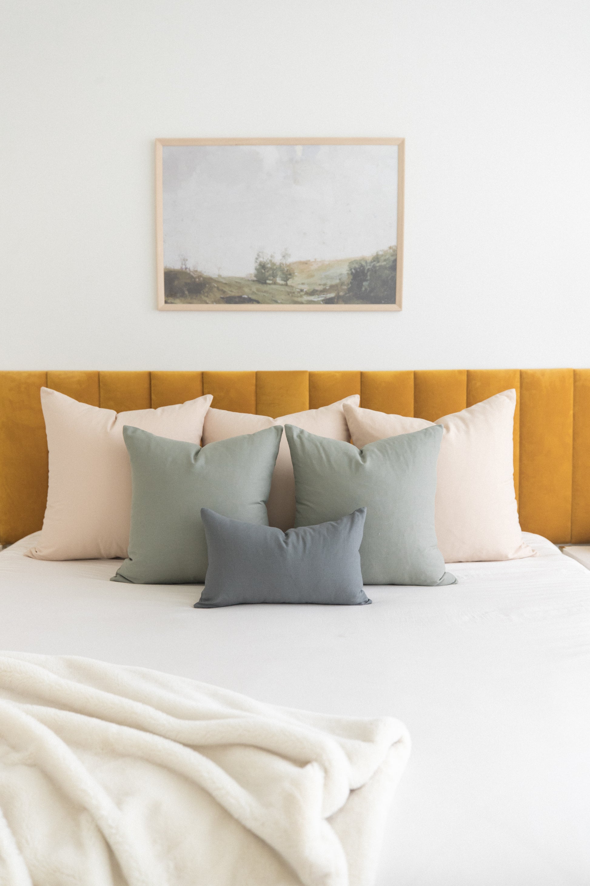 Beige and sage green throw pillows arranged on a bed with mustard yellow headboard and framed landscape artwork above.