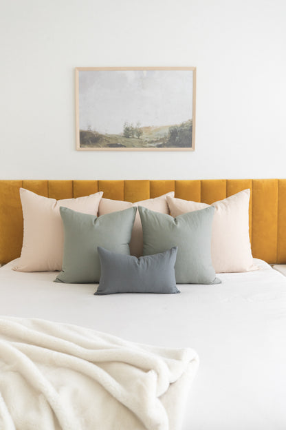 Beige and sage green throw pillows arranged on a bed with mustard yellow headboard and framed landscape artwork above.