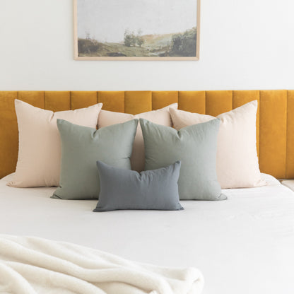 Collection of five solid-colored throw pillows in cream, sage green, and slate blue arranged on a bed with mustard headboard.