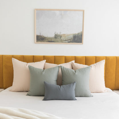 Light wood framed landscape artwork above bed with sage green and blue pillows on cream and golden upholstered headboard.