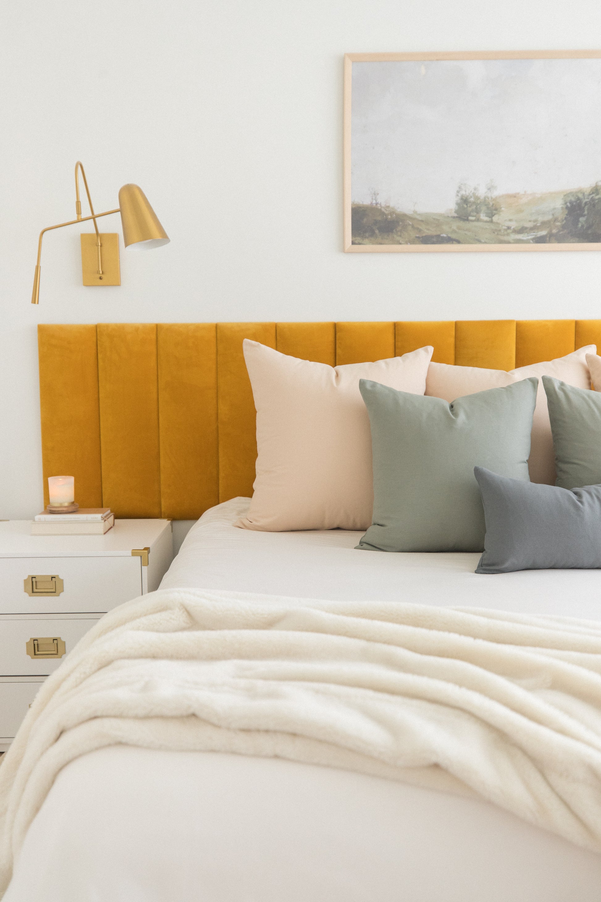 Bed with mustard upholstered headboard, cream and sage pillows, white bedding, gold swing lamp, and framed landscape artwork.