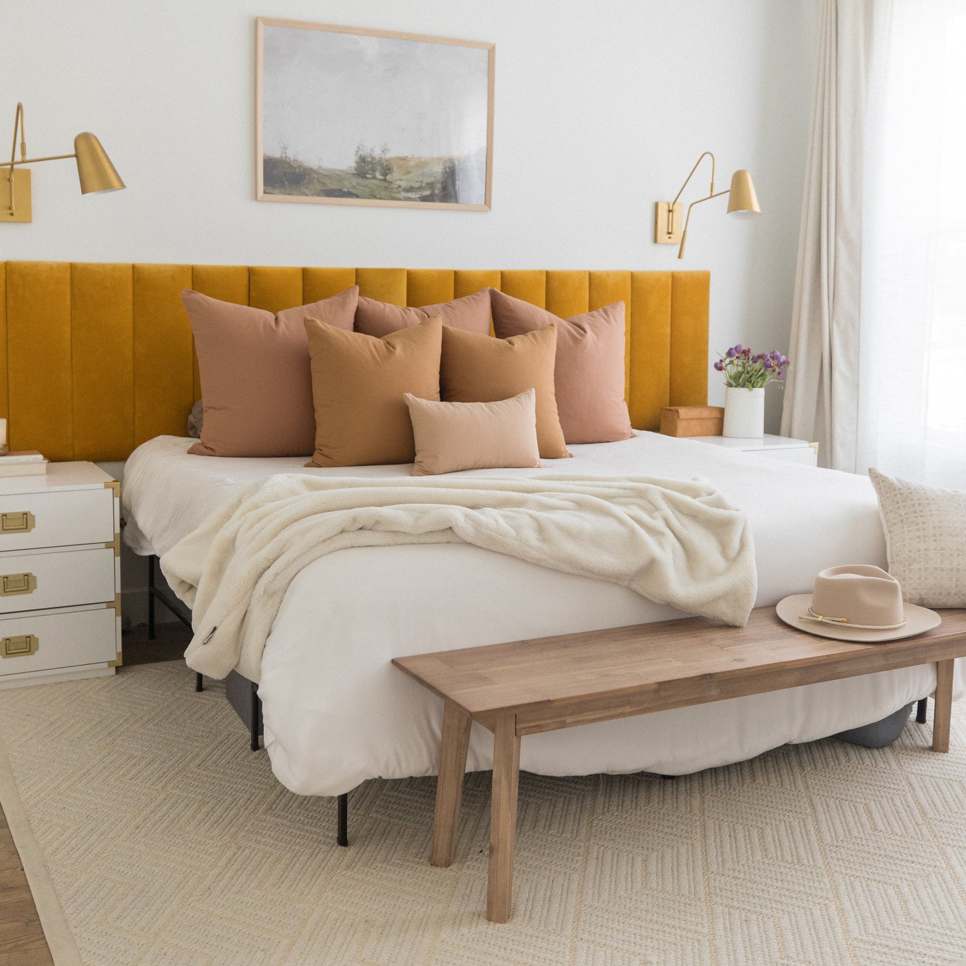 Bedroom with yellow-gold upholstered headboard, terracotta and tan throw pillows, cream bedding, and wooden bench at foot of 