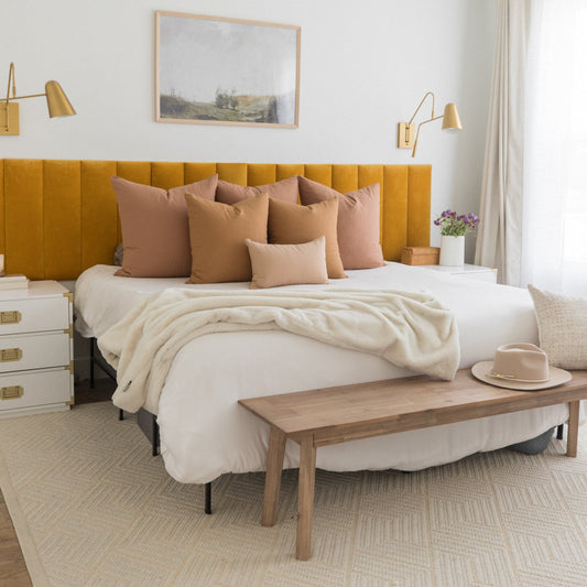Bedroom with yellow-gold upholstered headboard, terracotta and tan throw pillows, cream bedding, and wooden bench at foot of 
