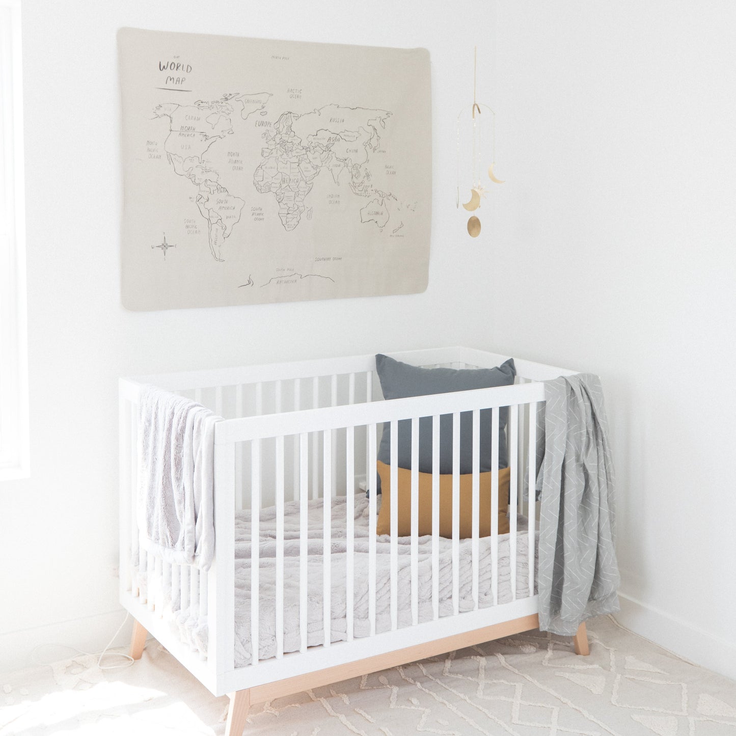 Beige linen world map wall hanging with hand-drawn country labels displayed above white crib with wooden legs.