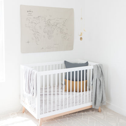 Beige linen world map wall hanging with hand-drawn country labels displayed above white crib with wooden legs.